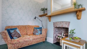 A vintage-style living room with a patterned sofa, a fireplace, a mirror, and a small table with a basket of apples. - 1 bedroom accommodation in Paignton (Paignton)