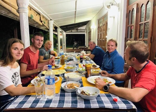 Un groupe de personnes est assis à une table à l’extérieur, recouverte d’une nappe à carreaux, et est entouré de différents plats et boissons.