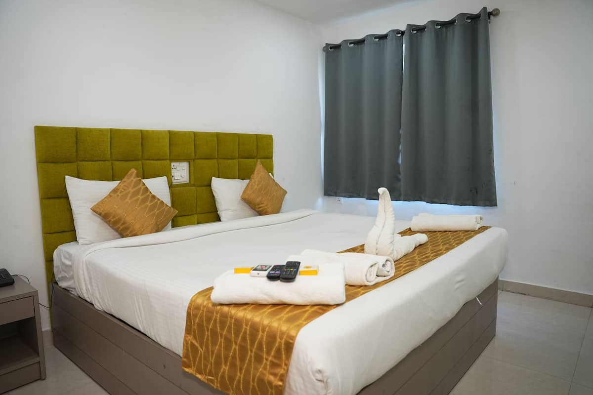 Habitación de hotel con cabecera verde, ropa de cama blanca, almohada decorativa marrón, cama con toallas y controles remotos, y ventana con cortinas oscuras.