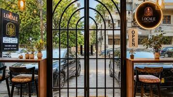 L’entrée d’un café, avec un sol à motifs géométriques, deux tables en bois accompagnées de chaises, et une vue sur les voitures garées et les bâtiments à l’extérieur.