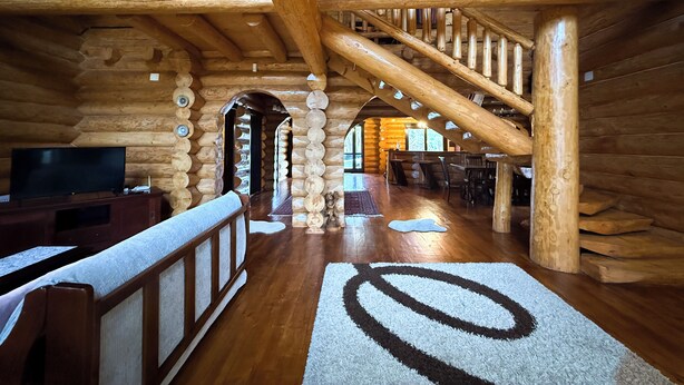 Interior de una cabaña de troncos con escalera de madera, un televisor sobre un mueble de madera y un tapete con diseño circular.