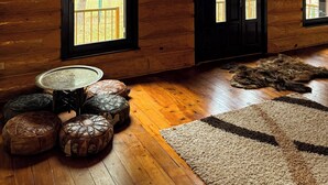 Interior de una cabaña de troncos con paredes de madera, techo alto y ventanas grandes. Seis otomanes de cuero, una mesa redonda y una alfombra mullida sobre el piso de madera.