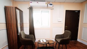Una habitación con dos sillones, una mesita redonda, un armario, un ventanal con cortinas y un equipo de aire acondicionado.