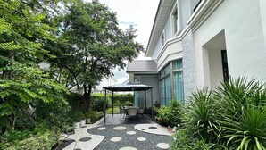 Un patio exterior moderno con una zona de estar cubierta, rodeado de abundante vegetación y un sendero con diseño.