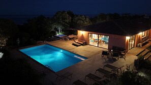 A swimming pool with lounge chairs and a patio area, surrounded by a house with a visible interior and a view of the sea. - Les Hauts de la Testa (Sainte-Lucie de Porto-Vecchio)