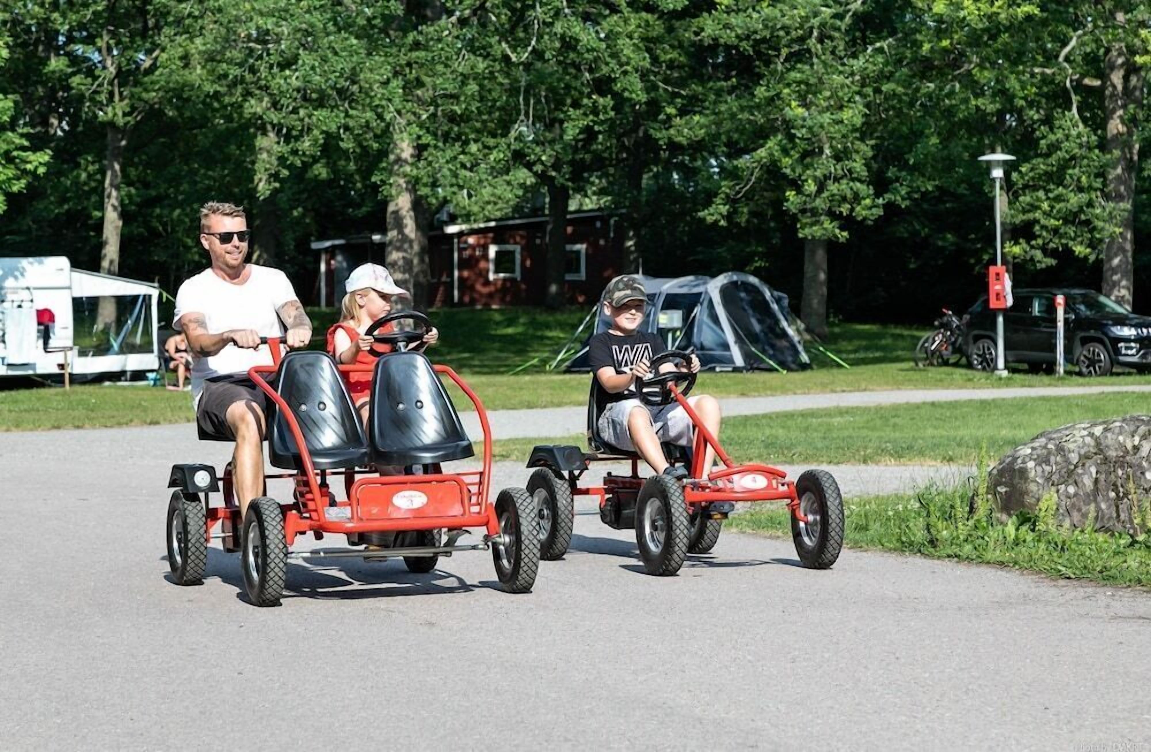 Three people on a red go-kart, a tent, and a parked car in a park.