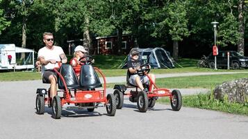 Three people on a red go-kart, a tent, and a parked car in a park.