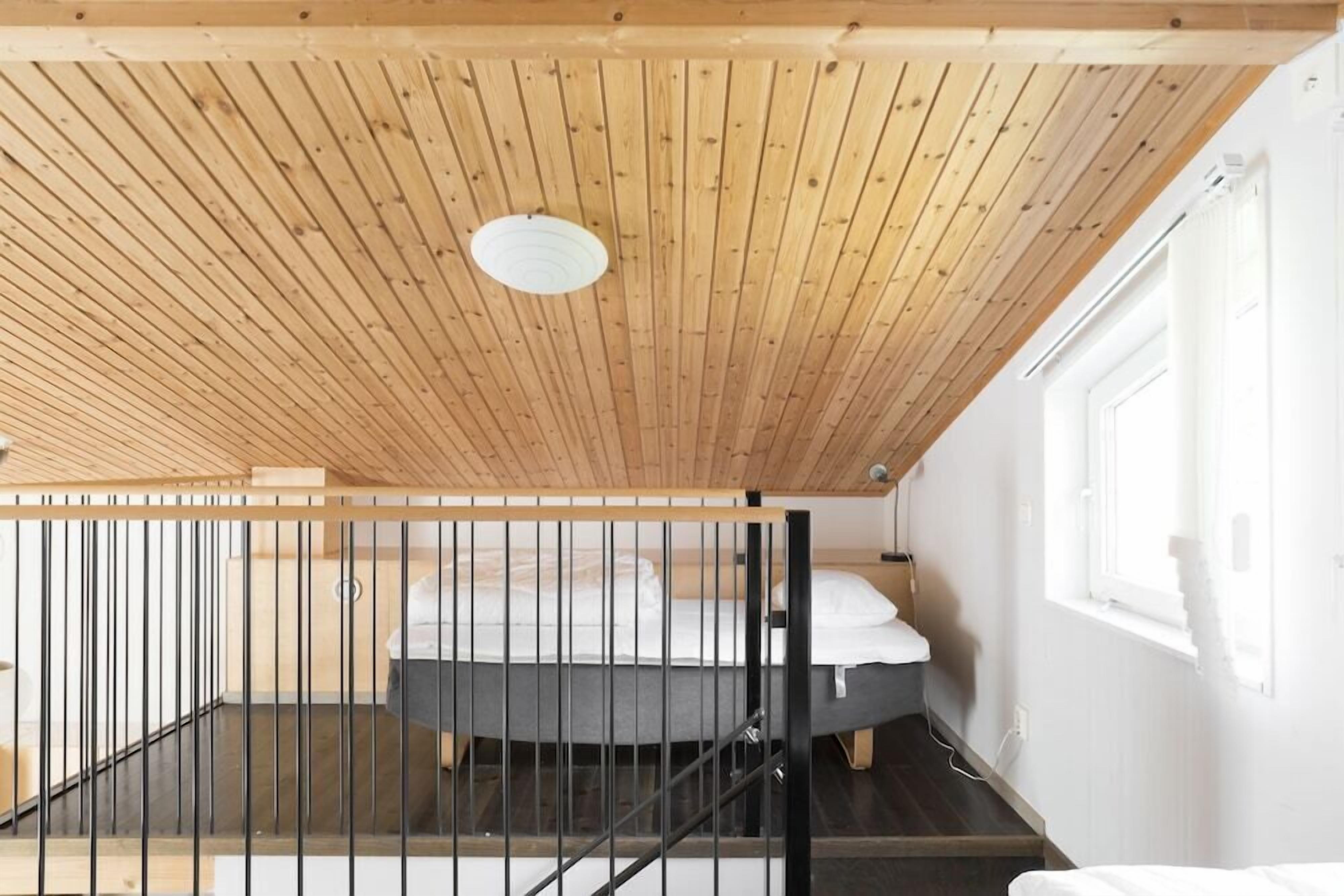 Ein Zimmer mit schrägen Wänden, einem Bett mit weißer Bettwäsche, einem Fenster und einer Treppe mit Geländer.