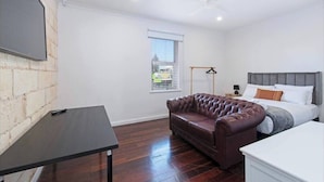 A bedroom with a bed, a brown leather sofa, a small table, and a window with blinds.