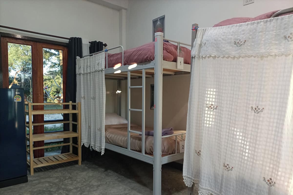 Une chambre avec un lit superposé, un réfrigérateur, une étagère en bois et une fenêtre avec des rideaux.