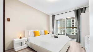 Ein modernes Hotelzimmer mit einem großen Bett, einem Nachttisch mit Lampe, einem Kleiderschrank und Blick auf die Stadt durch ein großes Fenster.