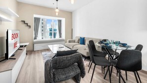 Ein modernes Wohnzimmer mit einer Couch, Couchtisch und einem Esstisch für sechs Personen. Ein Fernseher ist an der Wand montiert, und es führen Treppen in einen oberen Bereich.