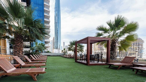 Eine Dachterrasse mit Holzliegen und einer Holzpergola, umgeben von Palmen und einer Stadtkulisse im Hintergrund.