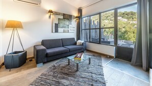 Ein modernes Wohnzimmer mit grauem Sofa, einem Glastisch, einem großen Fenster mit Blick auf eine Stadtlandschaft und einer Stehlampe.