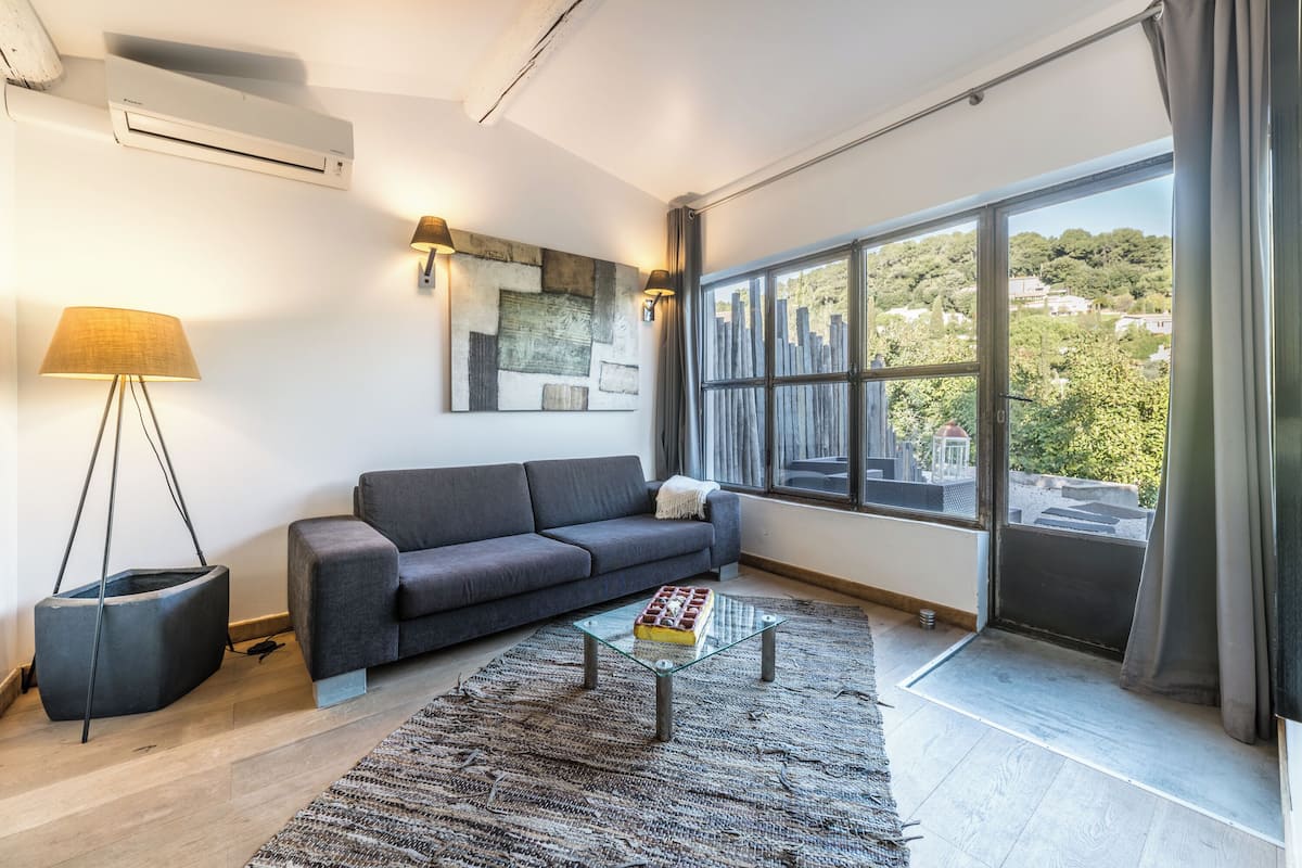 A modern living room with a grey sofa, a glass coffee table, a large window with a view of a cityscape, and a floor lamp.