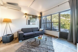 Una sala de estar moderna con un sofá gris, una mesa de centro de vidrio, un ventanal con vistas a un paisaje urbano y una lámpara de pie.