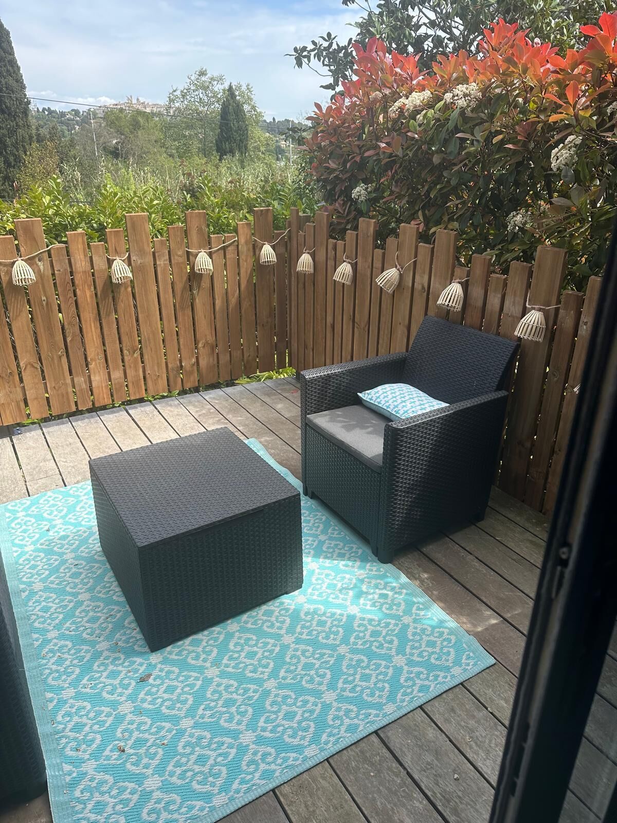 A patio with two wicker chairs, a matching ottoman, and a wooden fence with hanging lights.