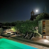Un área de piscina nocturna con sillones de descanso, un muro de piedra y una iglesia con campanario al fondo.