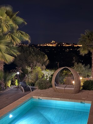 A swimming pool with a wooden deck, a wicker chair, and a view of a lit-up building in the background. - Villa 45 (La Colle-sur-Loup)