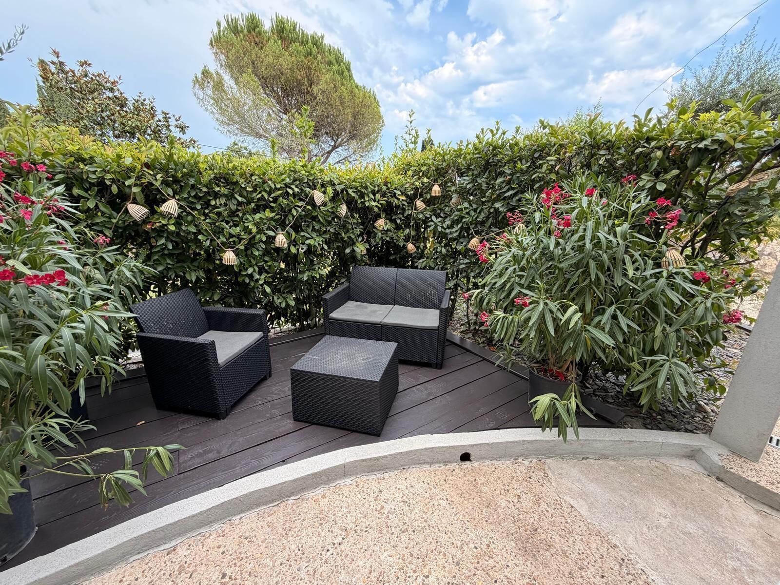 Um pátio externo com mobiliário de vime e piso de pedra, cercado por vegetação exuberante e plantas floridas.