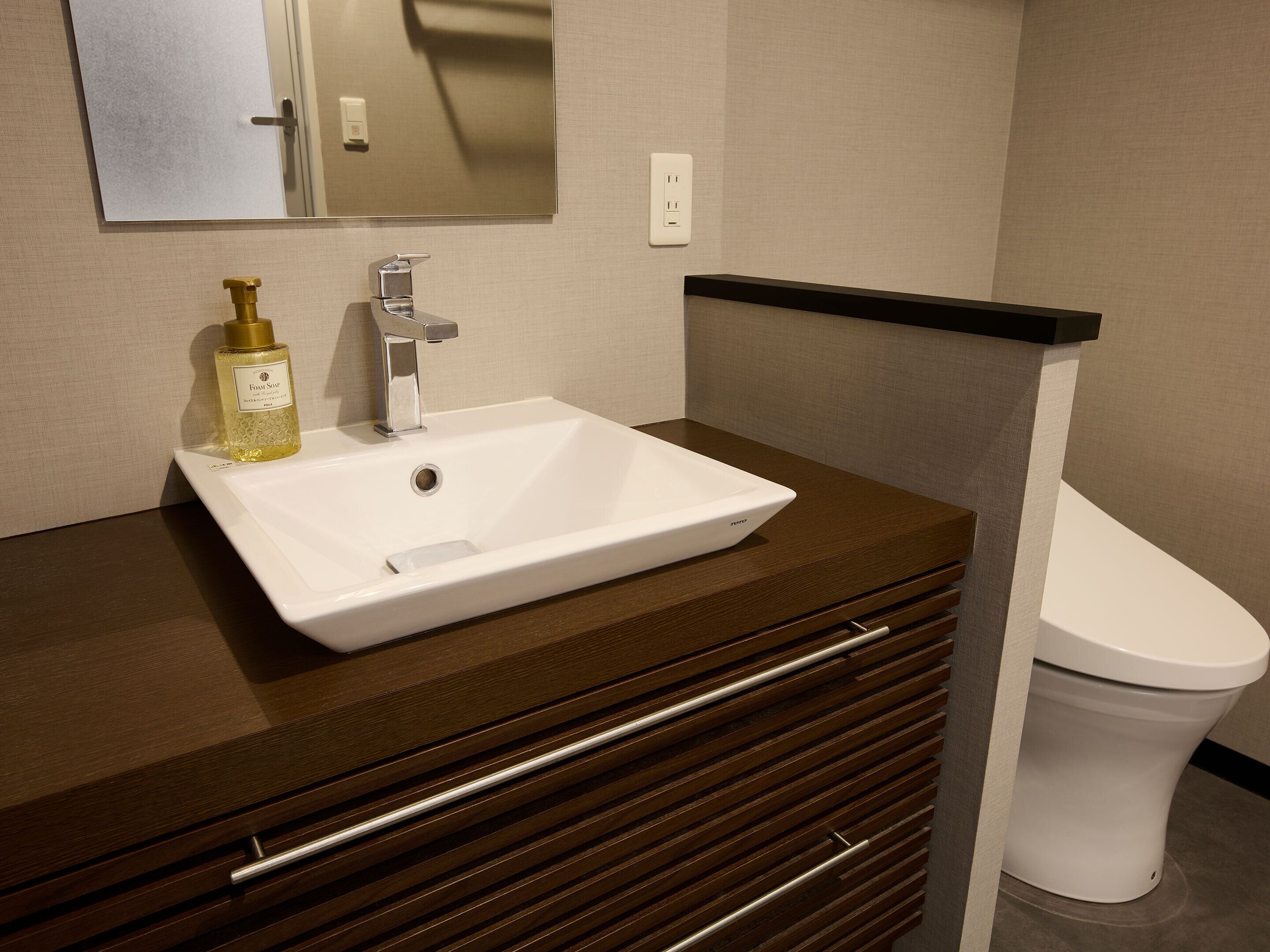 A bathroom with a white sink, a mirror, a bottle of hand soap, and a toilet.