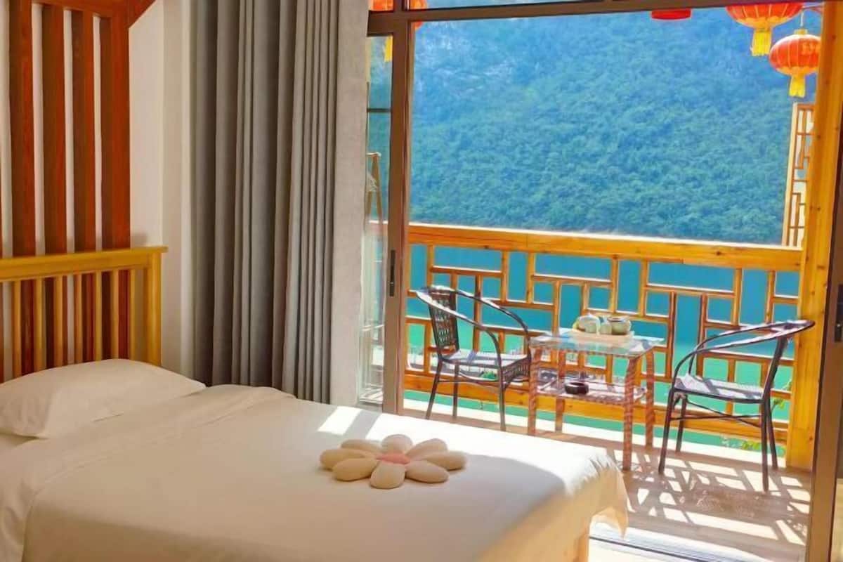 A hotel room with a bed, a balcony with a table and chairs, and a view of a forested mountain.