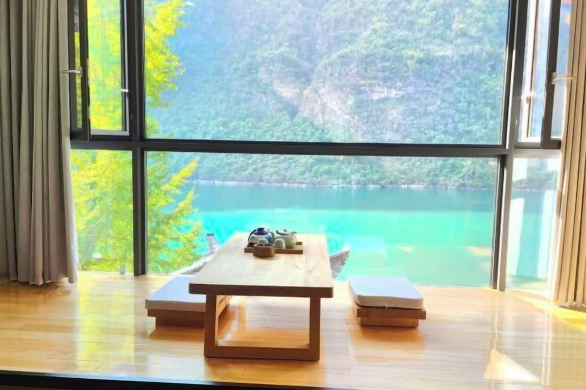 A room with a wooden table and two cushions, large windows with a view of a lake and forest.