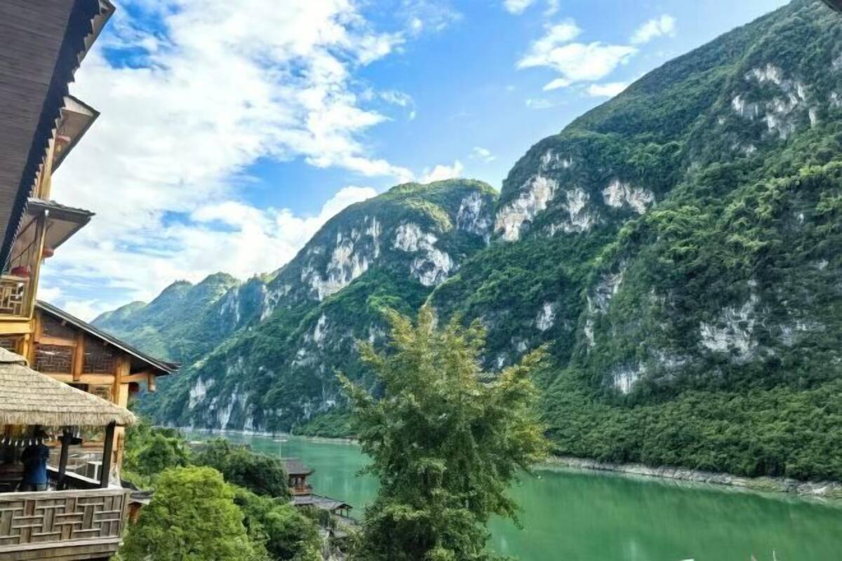 A traditional wooden building with a thatched roof overlooking a green lake and mountains.