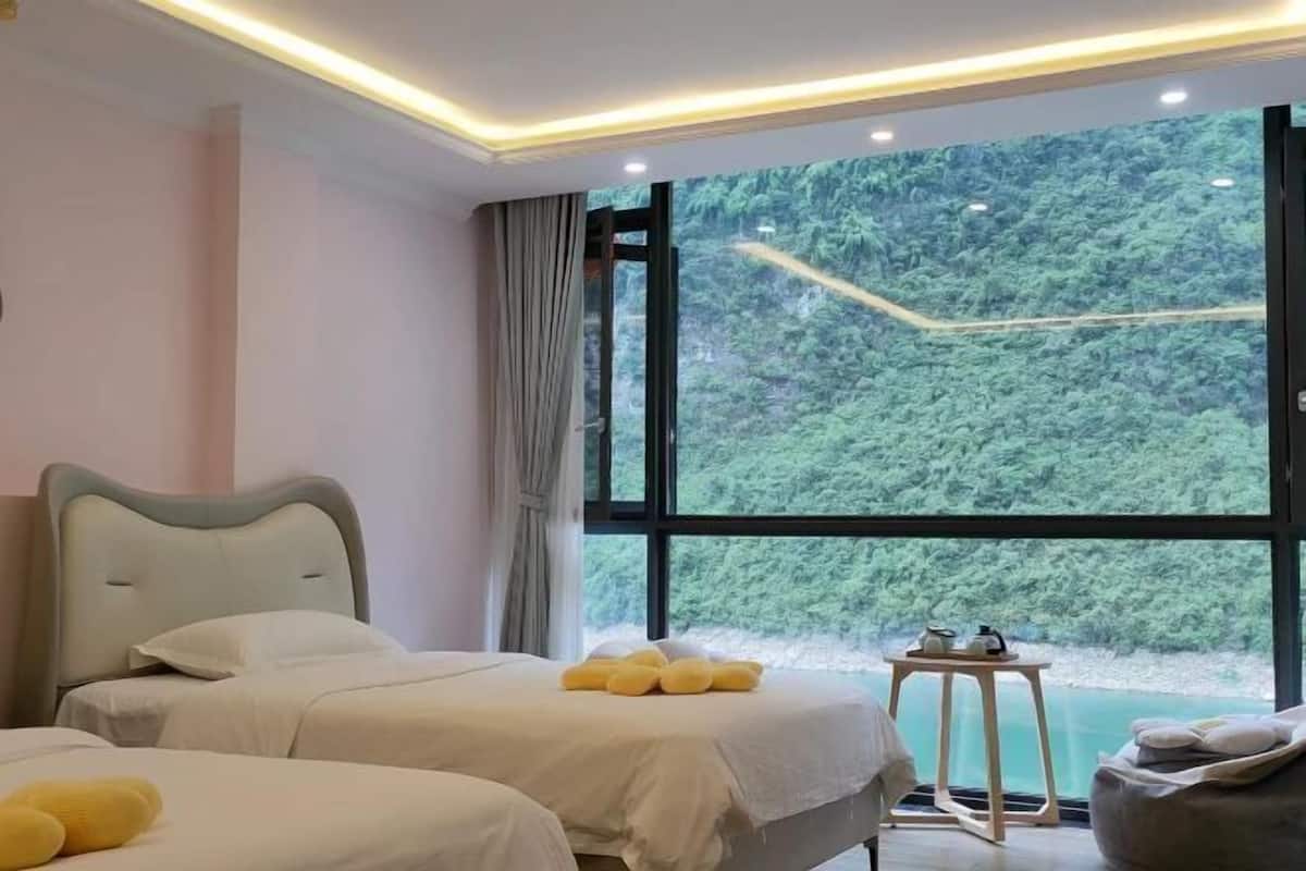 A bedroom with a large window, a bed, a bedside table, and a view of a forest.