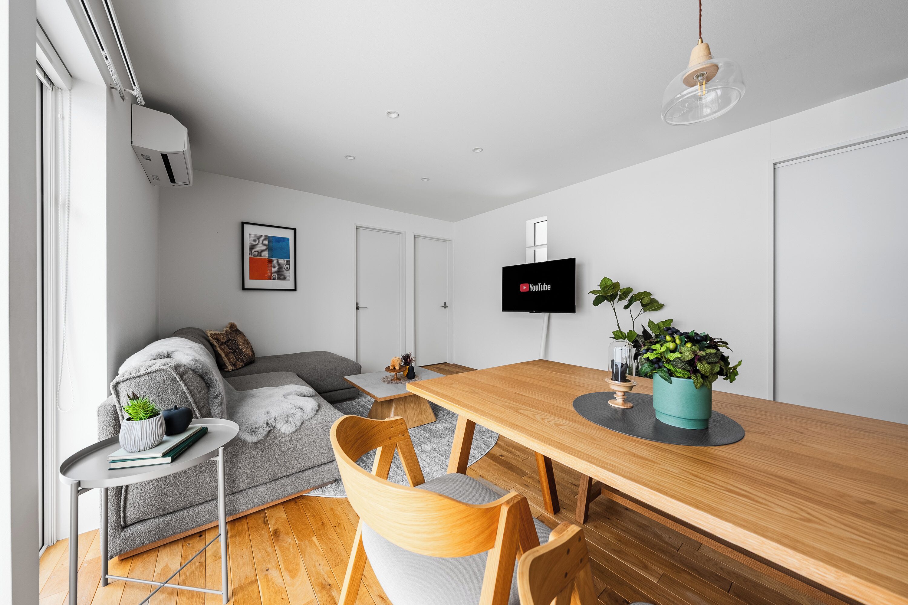 A modern living room with a grey sofa, wooden dining table, and chairs. A flat-screen TV is mounted on the wall, and there is a potted plant on the table.