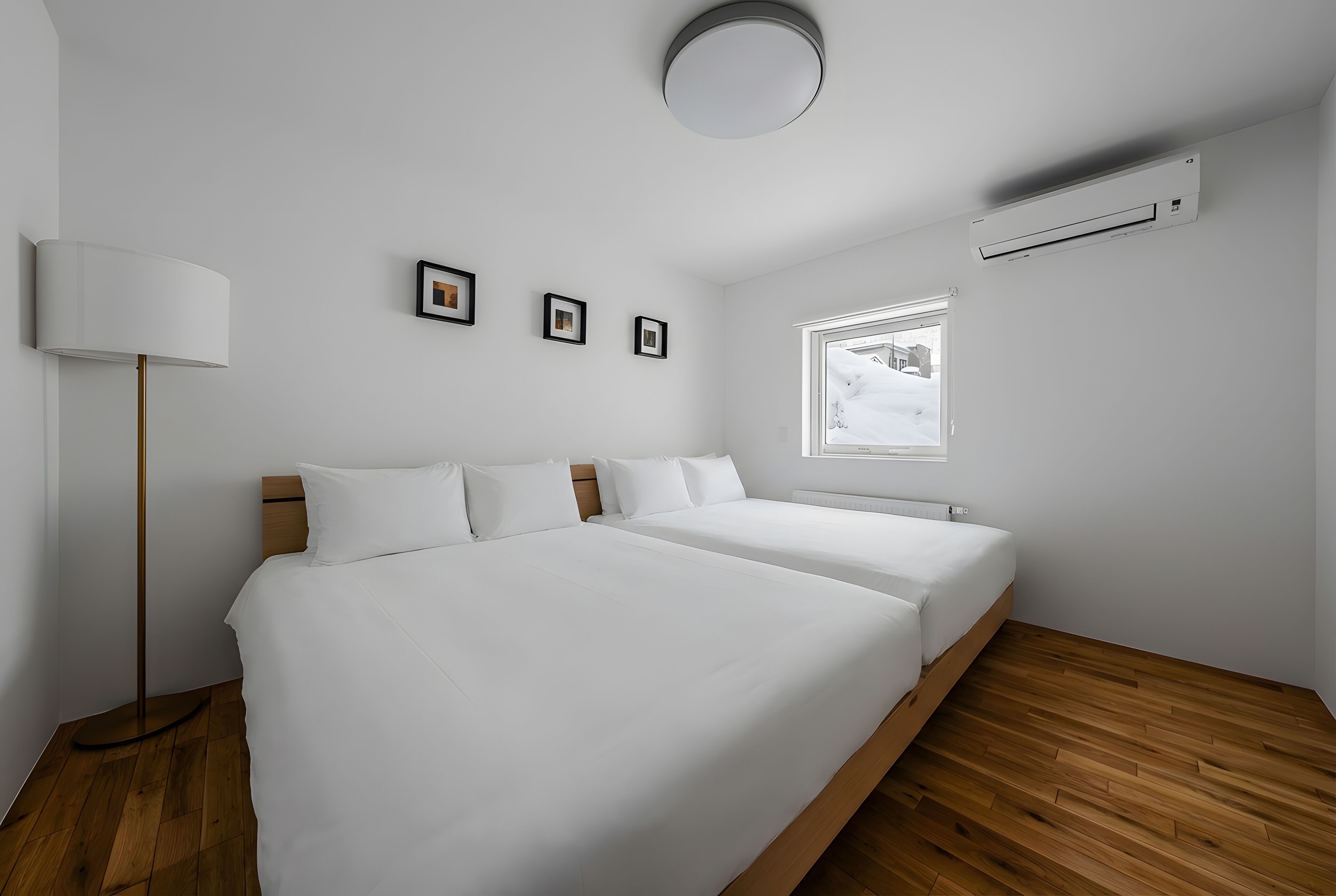 A modern bedroom with two beds, a standing lamp, a window with a view, and an air conditioning unit.