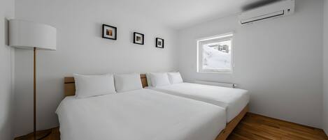 A modern bedroom with two beds, a standing lamp, a window with a view, and an air conditioning unit.
