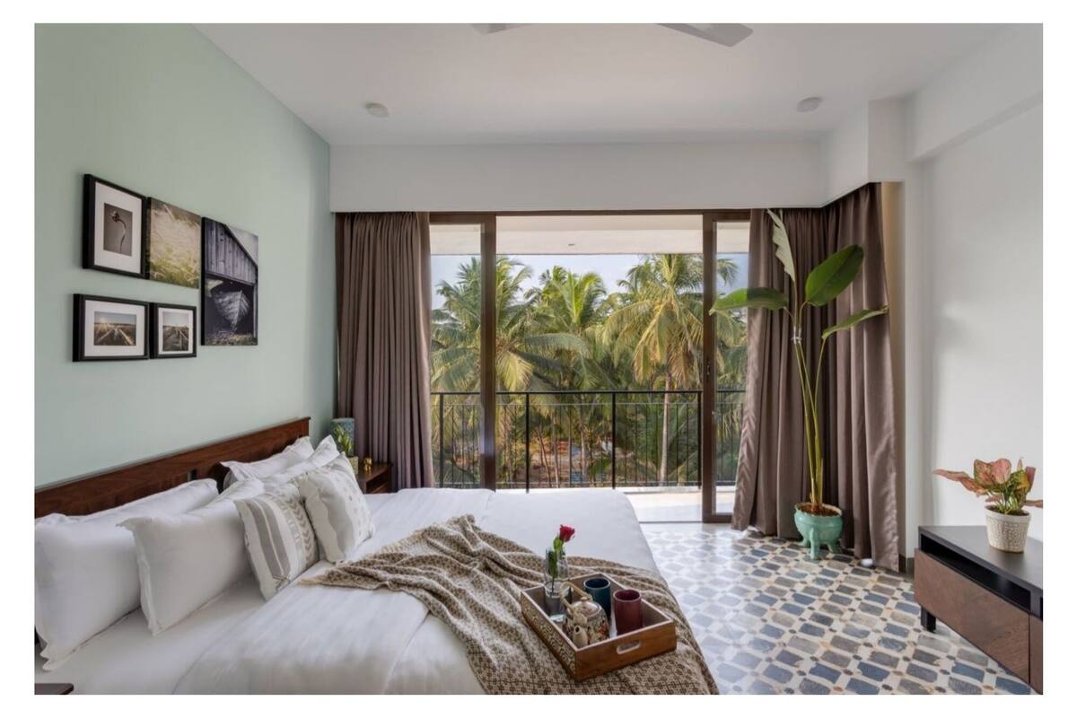 A hotel room with a large bed, a balcony with a view of palm trees, and framed pictures on the wall.