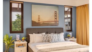 Et soveværelse med en stor seng, en sengegavl, et natbord, en lampe, et indrammet billede af Golden Gate Bridge og et vindue med gardiner.