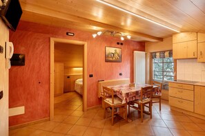 Un intérieur en bois comprenant une table à manger, des chaises, un coin cuisine et une chambre à coucher visible à travers une porte ouverte.