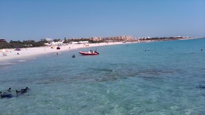 A beach with clear turquoise water, a red boat, and a sandy shore with umbrellas. - VILLA L'OLIVIER (Kelibia)