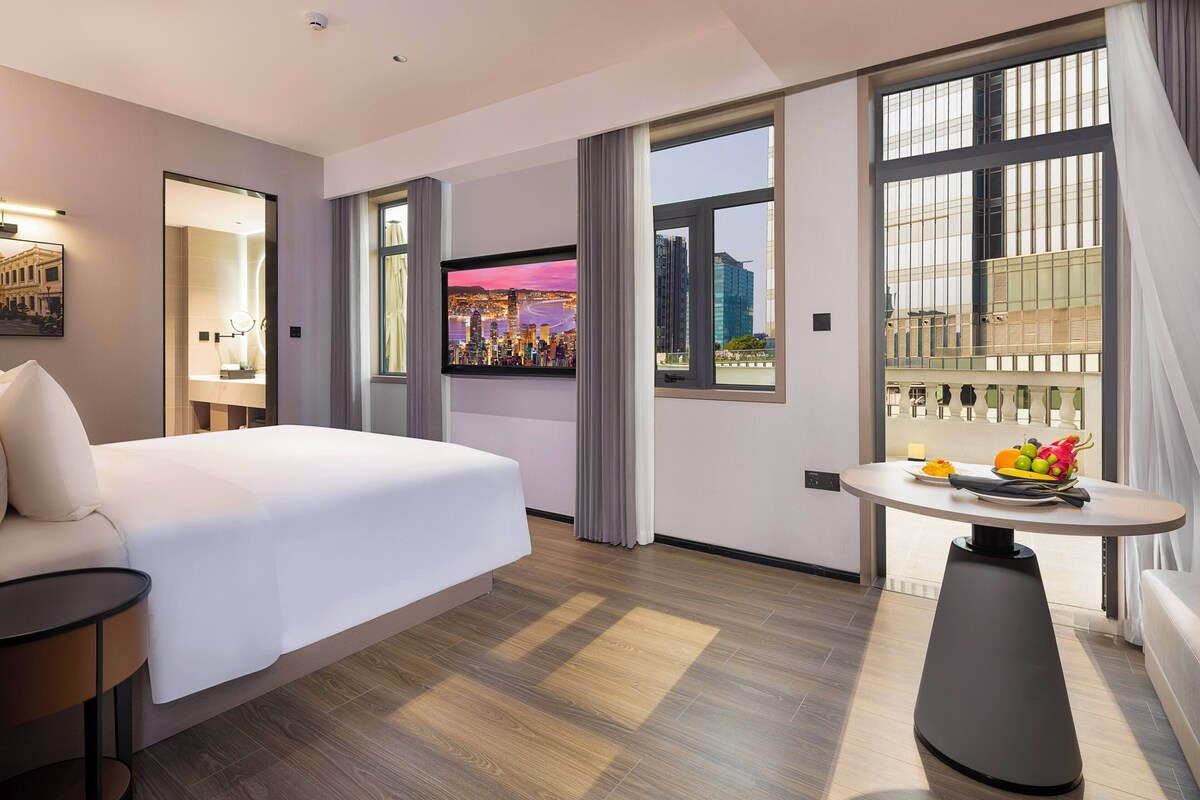 A hotel room with a large bed, a round table with fruit, a cityscape view, and a modern design.