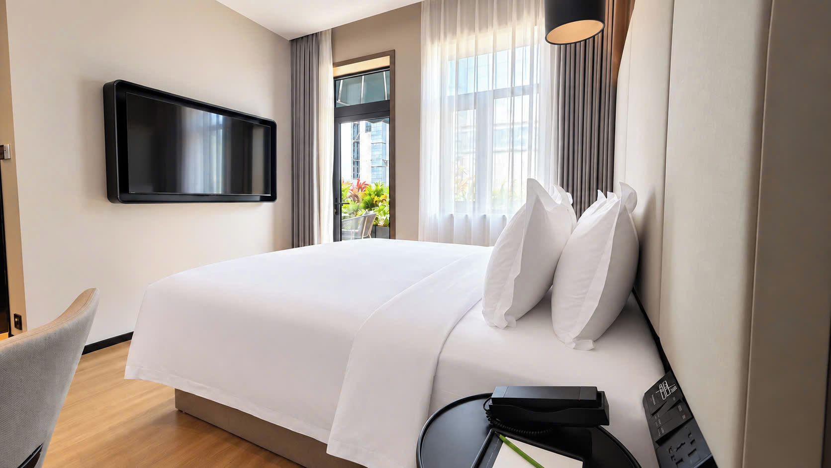 Habitación de hotel con cama, televisor montado en la pared, escritorio con silla y ventana con cortinas.
