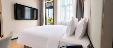 Habitación de hotel con cama, televisor montado en la pared, escritorio con silla y ventana con cortinas.