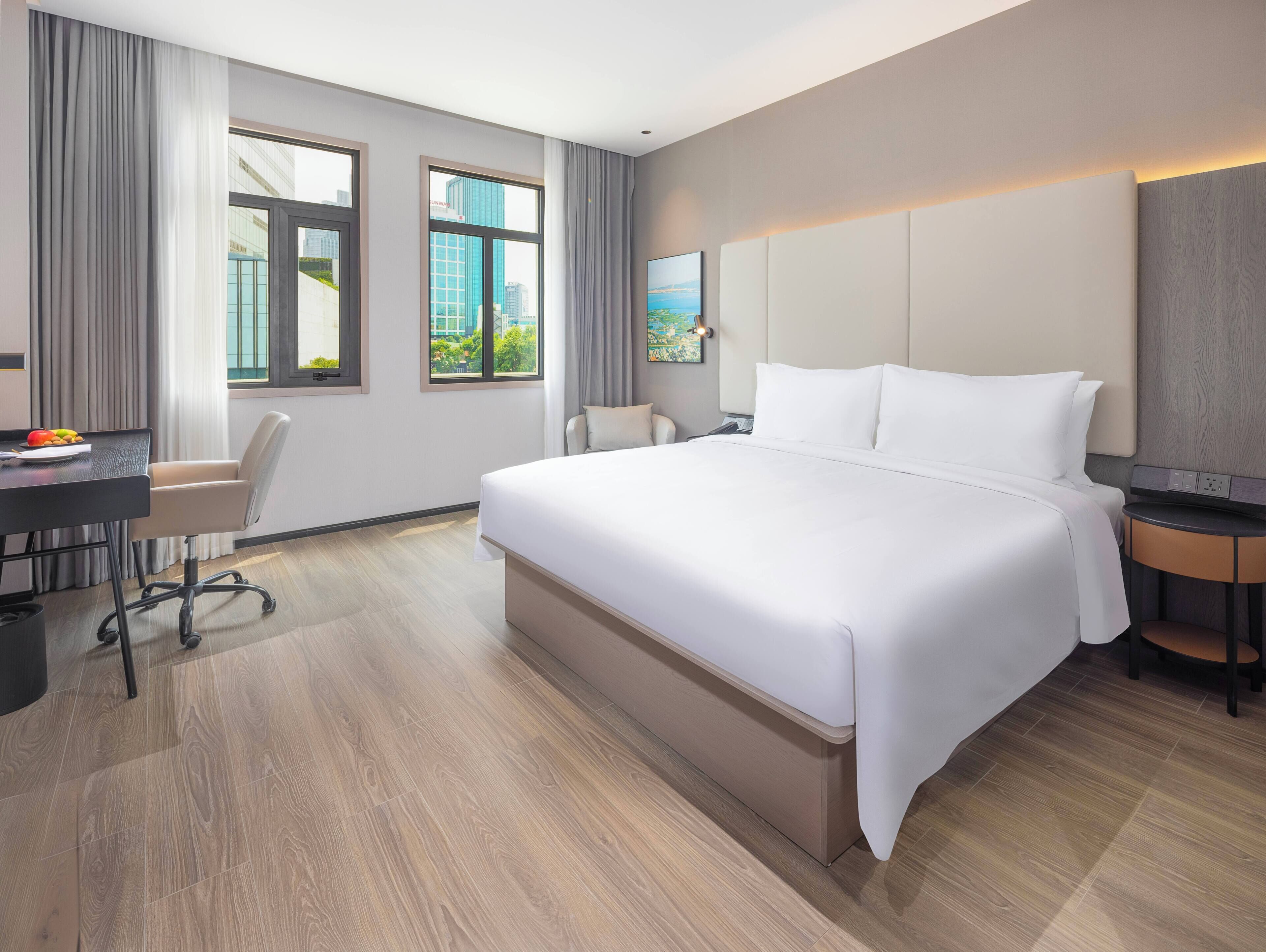 A hotel room with a large bed, a desk with a chair, and a view of buildings through the windows.