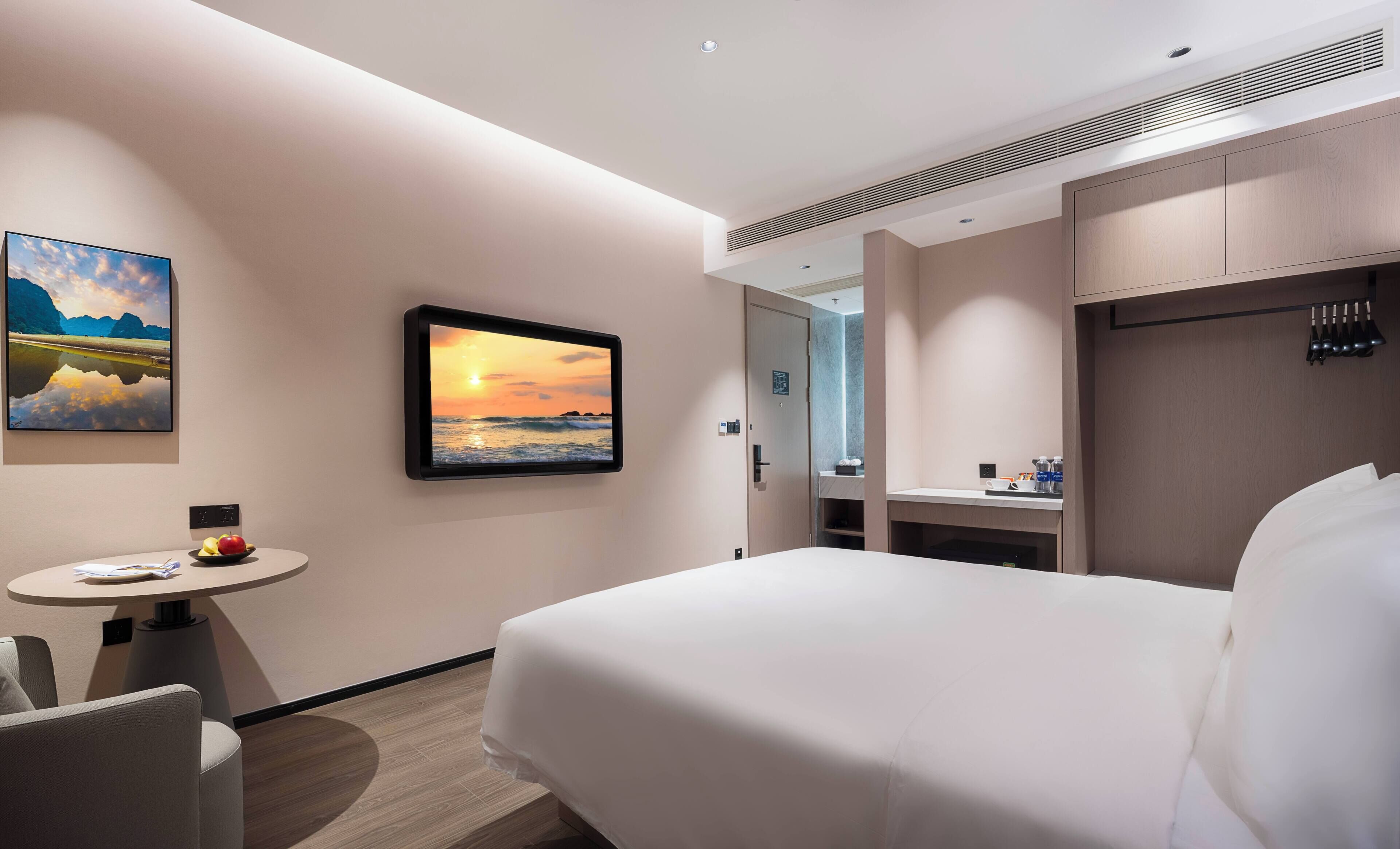 A hotel room with a bed, a desk, a chair, a small table with fruit, a painting of a sunset, and a mirror.