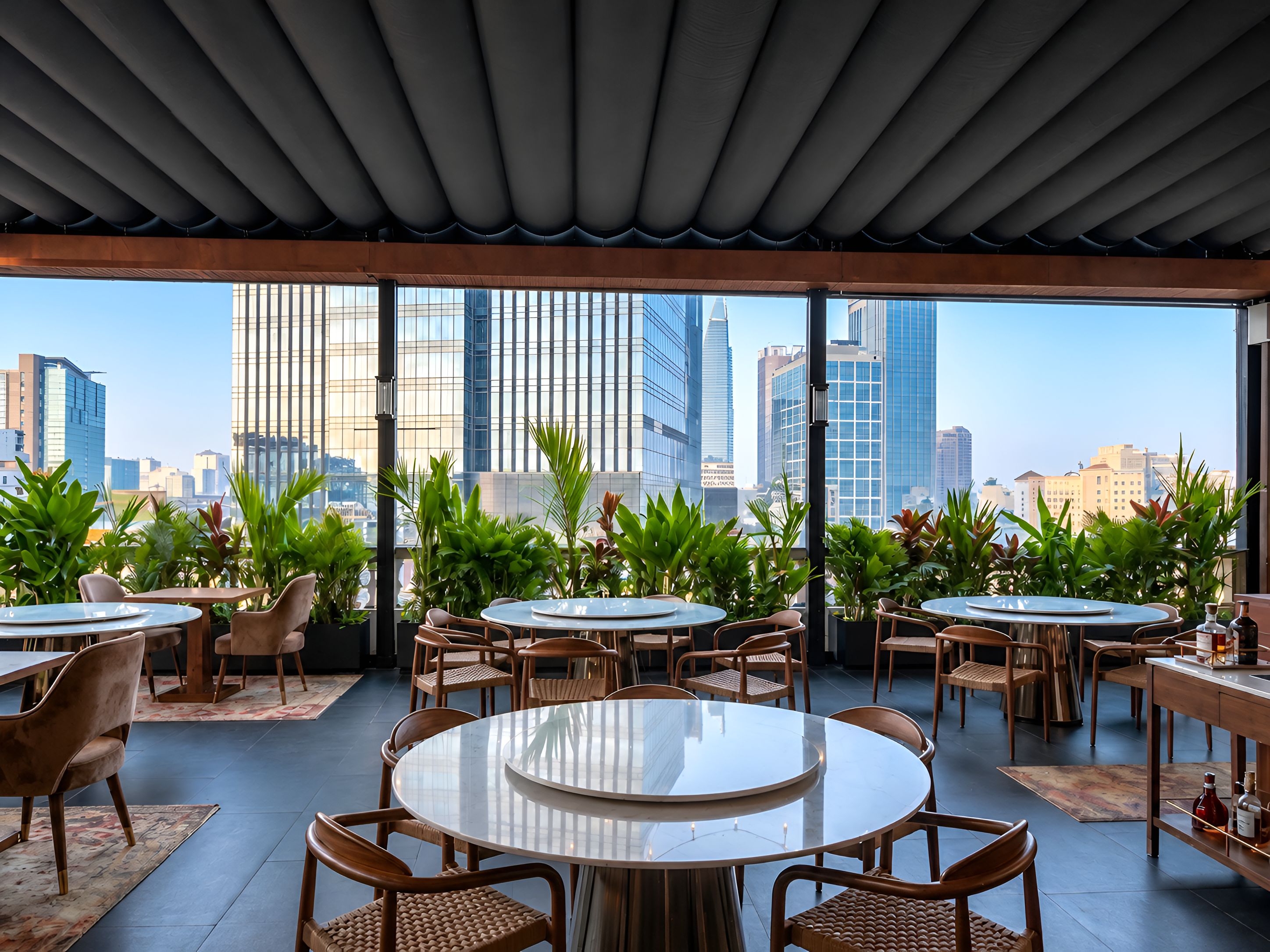 Un espace de restauration aménagé sur le toit, avec des tables et des chaises rondes, entouré de plantes en pot, et offrant une vue panoramique sur la ville.