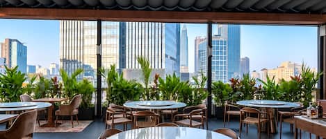 A rooftop dining area with round tables and chairs, surrounded by potted plants, and offering a city skyline view.