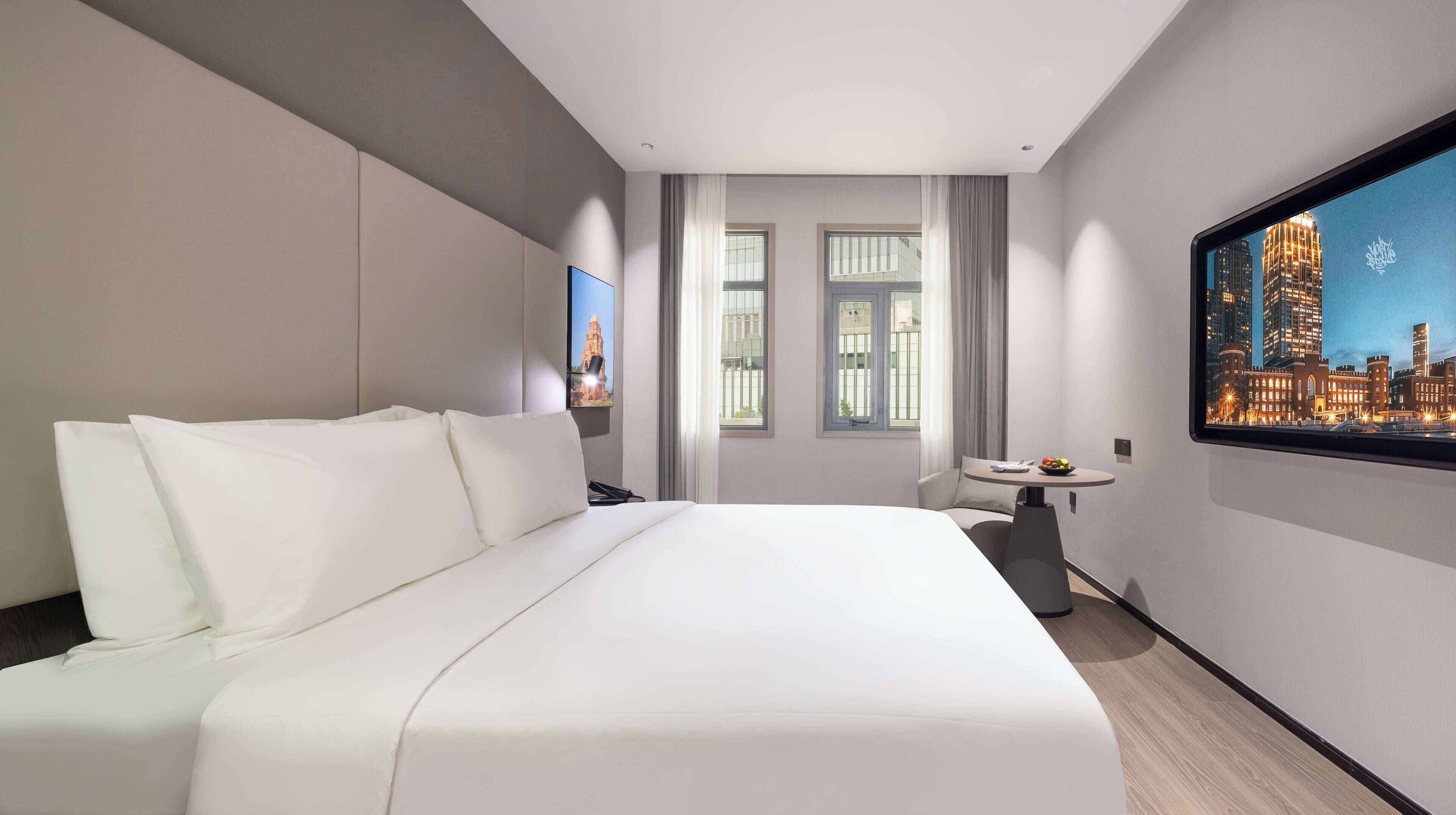 Ett hotellrum med en stor säng, ett litet bord med en fruktskål, en tv som visar en stadsbild och ett fönster med gardiner.