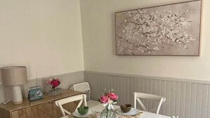 A dining area with a white table and chairs, a decorative vase with flowers, a wall-mounted artwork, and a sideboard with a lamp and a vintage radio.