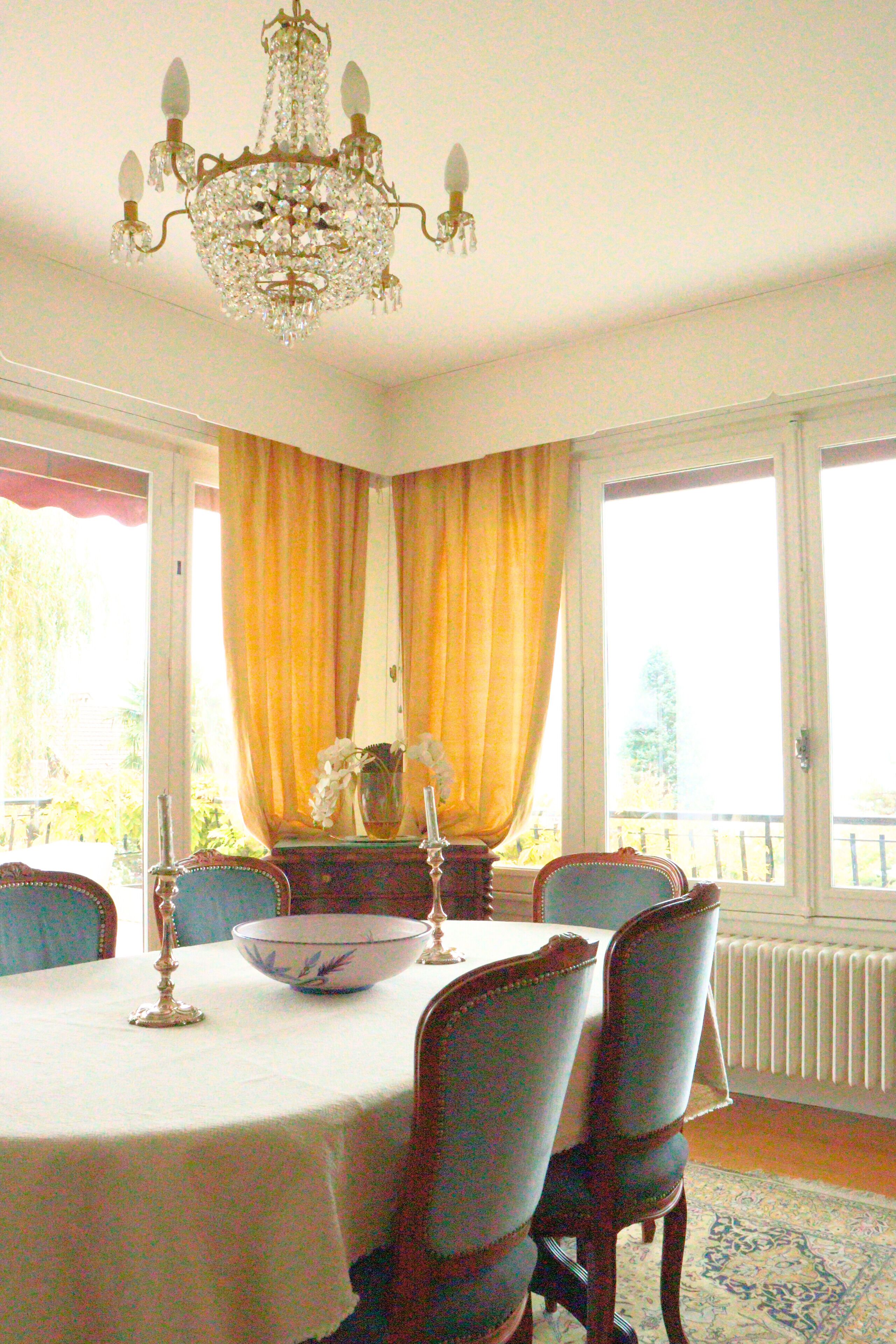 Ein Esszimmer mit einer Kandelaber, einem Tisch mit Schüssel und Kerzen, Stühlen und Blick durch große, mit Vorhängen drapierten Fenster.