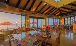 A spacious dining area with multiple tables and chairs, large windows offering a view of the sea, and ceiling fans. - Luxury 2BR Sanctuary Retreat Overlooking Banderas Bay (Puerto Vallarta)
