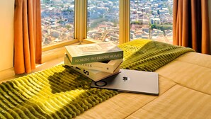 Een kamer met een groot raam dat uitzicht biedt op de stad, een bed met een groene deken en een laptop op het bed.