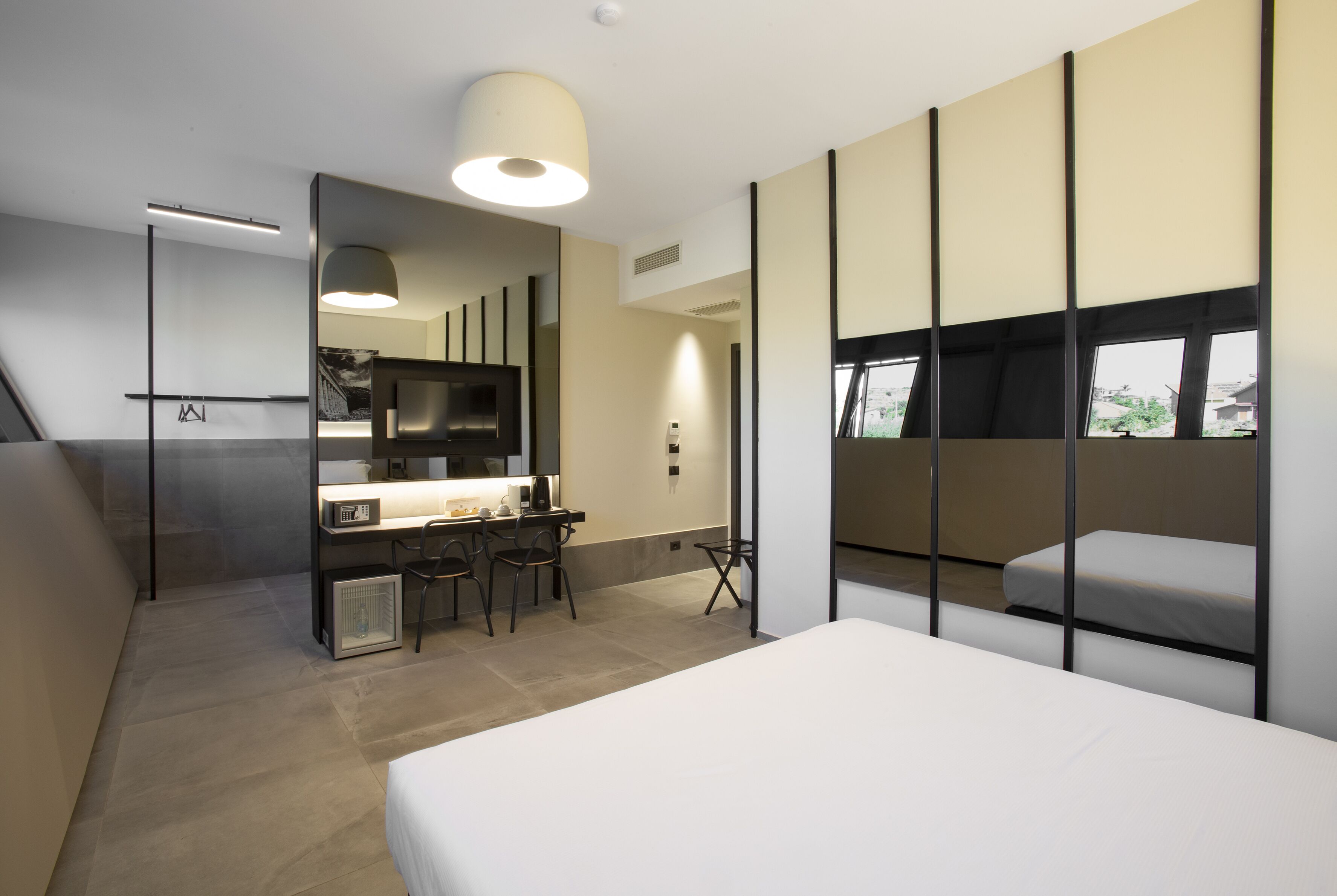 Una camera d'albergo moderna con un letto, una scrivania con sedie, una TV e una grande finestra.