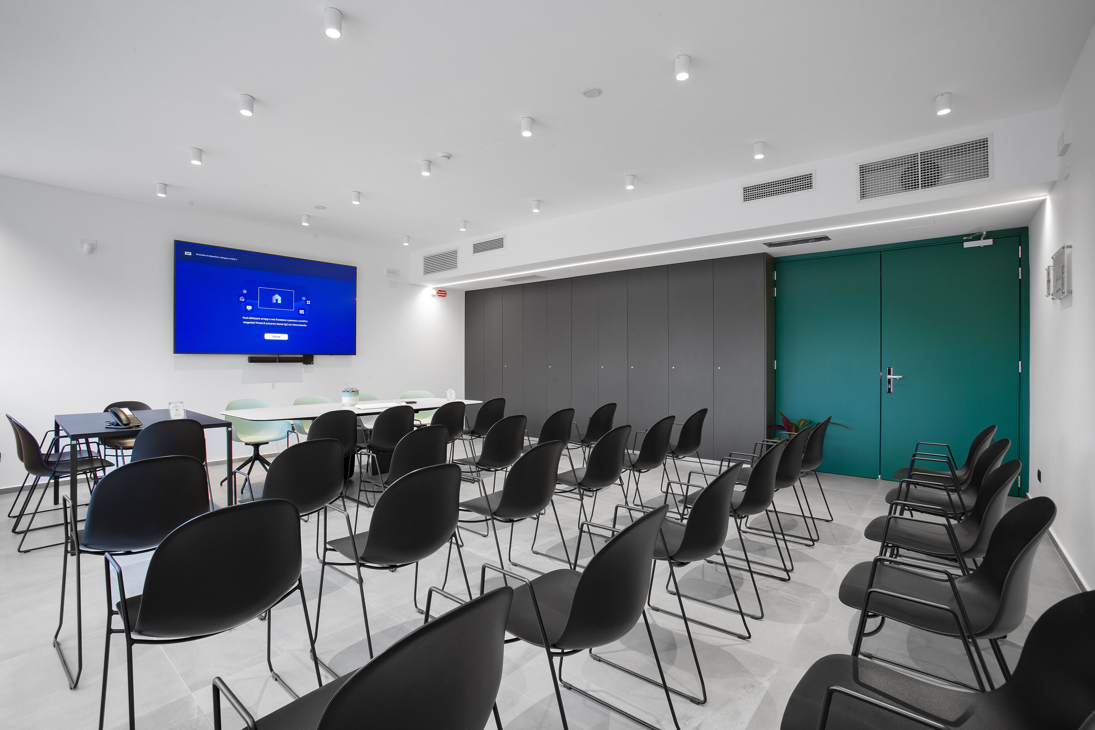 Un salón de conferencias con filas de sillas, una mesa, un televisor de pantalla plana y una puerta color turquesa.